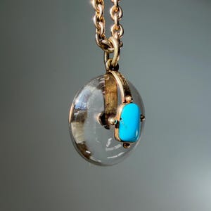 Op de afbeelding: Een goudkleurige ketting met een ronde, transparante hanger en een rechthoekige turkooizen steen. De hanger hangt aan een fijne ketting. De turkooizen steen zorgt voor een opvallend contrast. Een klassiek en elegant sieraad.