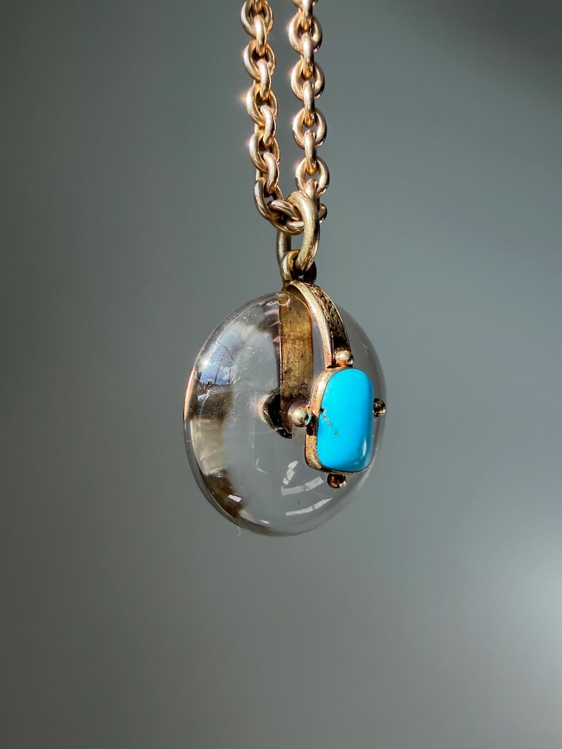 Op de afbeelding: Een goudkleurige ketting met een ronde, transparante hanger en een rechthoekige turkooizen steen. De hanger hangt aan een fijne ketting. De turkooizen steen zorgt voor een opvallend contrast. Een klassiek en elegant sieraad.