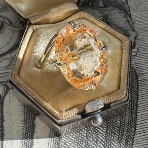 May include: A vintage ring with a large, oval-shaped, light orange gemstone set in a gold band. The ring is in a small, hexagonal, velvet-lined box with the inscription "Ellis Jewellers Toronto".