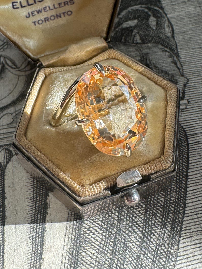 May include: A vintage ring with a large, oval-shaped, light orange gemstone set in a gold band. The ring is in a small, hexagonal, velvet-lined box with the inscription "Ellis Jewellers Toronto".