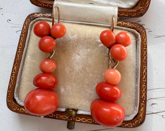 May include: A pair of coral earrings with a drop design. The earrings are made of multiple coral beads in shades of orange and red, and are suspended from gold hooks.