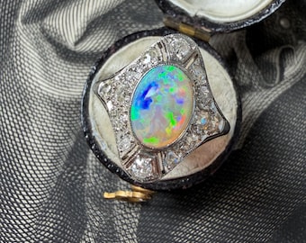 Puede incluir: Un anillo antiguo con una ópalo ovalado central que muestra colores iridiscentes de azul, verde y naranja, rodeado por un halo de pequeñas gemas transparentes. El anillo está engastado en una montura de plata u oro blanco y presentado en un joyero vintage.