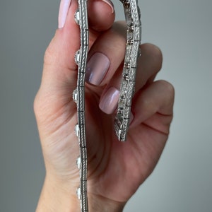 K&ouml;nnte beinhalten: Ein silbernes Armband mit einem detaillierten Design und mehreren kleinen Steinen. Das Armband wird in einer Hand gehalten.