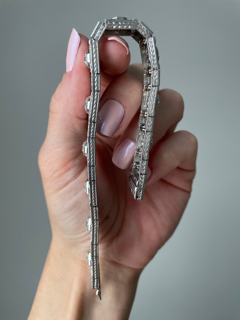 K&ouml;nnte beinhalten: Ein silbernes Armband mit einem detaillierten Design und mehreren kleinen Steinen. Das Armband wird in einer Hand gehalten.