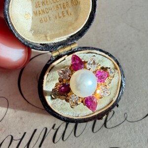Puede incluir: Un anillo de oro con una perla central rodeada de piedras preciosas rosas y claras. El anillo se presenta en un joyero vintage con el texto "Dickinson & Co. Jewellers Manchester Rd. Burley."