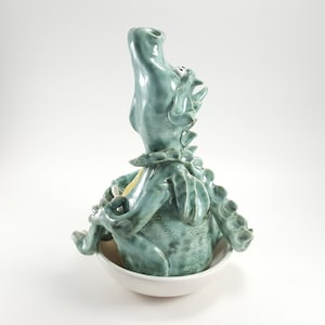 May include: A ceramic dragon figurine with a light blue glaze, sitting in a white bowl. The dragon has its head tilted back and its mouth open, as if it is roaring.