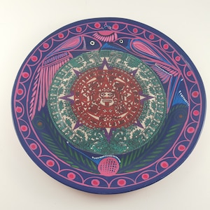 May include: A blue ceramic plate with a colorful design featuring a central circle with a red and white sun symbol, surrounded by two pink birds with blue and green accents. The plate has a decorative border of pink and blue.