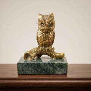 May include: A brass owl figurine perched on a textured branch, mounted on a rectangular green marble base. The owl features detailed feather patterns and large eyes, with the branch showing a hollow end on the left side.