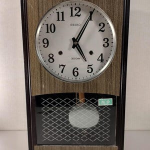 May include: Vintage Seiko 30-day wall clock with a light brown wood grain frame. The clock face is silver with black numerals and hands. The lower section has a dark glass panel with a grid pattern and a swinging pendulum.