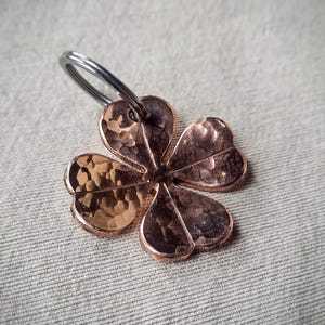 Pode incluir: Um chaveiro de trevo de quatro folhas cor de cobre com uma textura martelada. O trevo é preso a um anel prateado. O fundo é um tecido cinza claro.