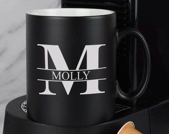 Könnte beinhalten: Schwarze Keramiktasse mit einem weißen Buchstaben "M" und dem Namen "MOLLY" darauf gedruckt. Die Tasse steht auf einer schwarzen Oberfläche.