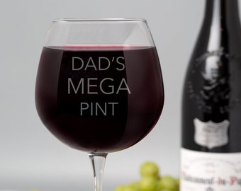 May include: A large, clear glass wine glass filled with red wine. The glass has the words "DAD'S MEGA PINT" etched on the side. The glass is sitting on a white surface with green and red grapes scattered around it.