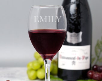 May include: A clear glass wine glass with the name "EMILY" etched on the side. The glass is filled with red wine and is sitting on a white surface with grapes in the background.