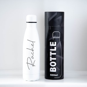 May include: White stainless steel water bottle with the name "Rachel" printed on it. The bottle is shown next to its black and white patterned carrying case. The case has the word "BOTTLE" printed on it and the text "500ml".