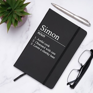 Pode incluir: Caderno preto com fecho de elástico branco. A capa frontal tem a palavra "Simon" escrita em branco, seguida da palavra "nome" e uma lista de três definições humorísticas.