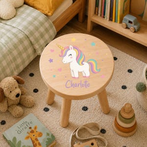 Op de afbeelding: Een ronde houten kruk met drie poten voorzien van een print van een witte eenhoorn met een veelkleurige manen en staart. Het ontwerp bevat kleine sterretjes en hartjes in diverse kleuren, met de naam Charlotte in paars schrift eronder.