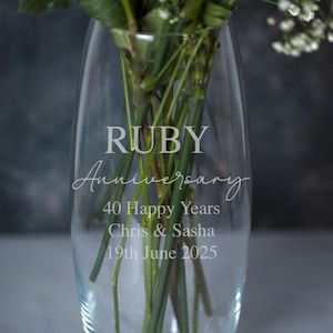 Può includere: Un vaso di vetro trasparente con le parole "RUBY Anniversary" e "40 Happy Years" incise sul davanti. Il vaso contiene steli verdi e fiori bianchi. I nomi "Chris & Sasha" e la data "19 giugno 2025" sono anch'essi incisi.
