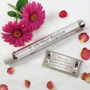 May include: A silver baptismal certificate holder with the inscription "On Your Baptism" and a cross. A small rectangular frame with engraved details and two pink gerbera daisies with rose petals are also present.