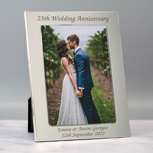 May include: Silver photo frame with a black mat and a wedding photo of a couple kissing. The frame is engraved with "25th Wedding Anniversary", "Emma & Aaron Georgio", and "12th September 2022".