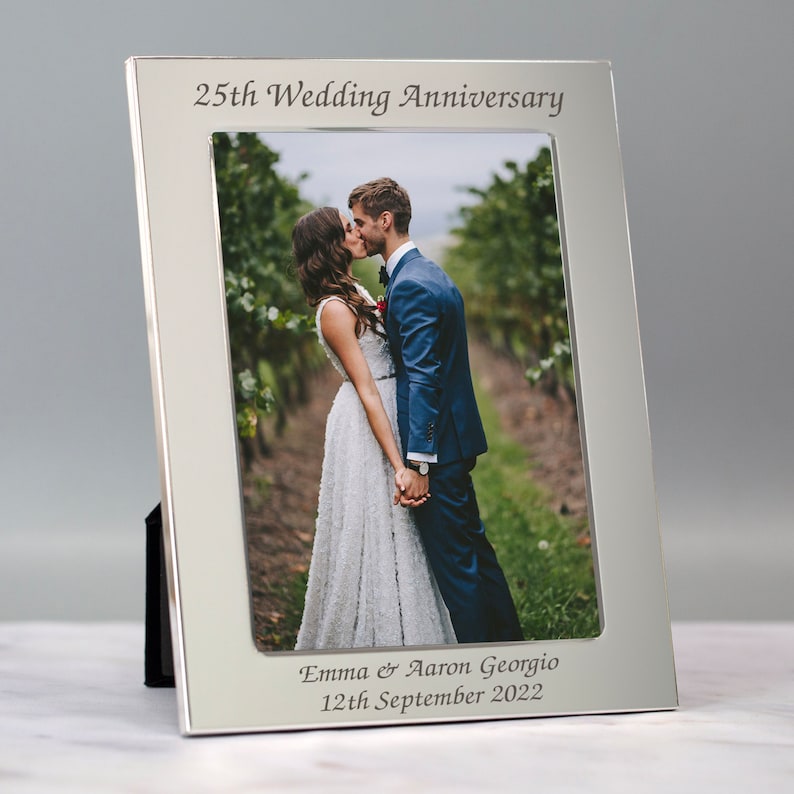 May include: Silver photo frame with a black mat and a wedding photo of a couple kissing. The frame is engraved with "25th Wedding Anniversary", "Emma & Aaron Georgio", and "12th September 2022".