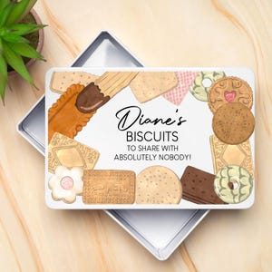 Op de afbeelding: Een witte, rechthoekige koektrommel met een verscheidenheid aan koekjes rond de randen. De trommel heeft de tekst "Diane's BISCUITS TO SHARE WITH ABSOLUTELY NOBODY!" in zwart script. De koekjes hebben verschillende vormen en kleuren.