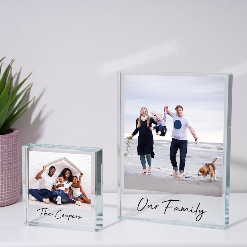 May include: Two glass photo frames with black text. The larger frame has a photo of a family of three on a beach with a dog. The text reads "Our Family". The smaller frame has a photo of a family of five sitting in front of a house. The text reads "The Coopers".
