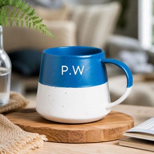 May include: A two-toned ceramic mug featuring a vibrant blue upper section and a white lower section with subtle speckles. The letters P.W are printed in white on the blue portion. The mug has a curved handle and a wide, stable base design.