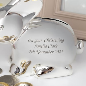 May include: Silver bunny piggy bank with a personalized inscription: "On your Christening Amelia Clark 7th November 2021".