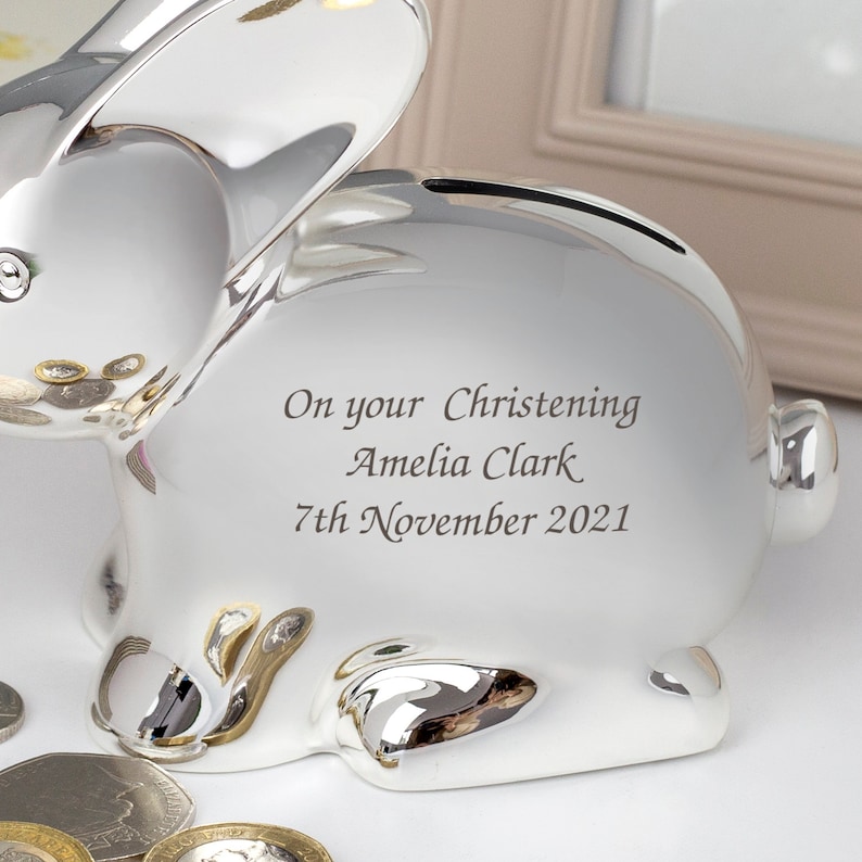 May include: Silver bunny piggy bank with a personalized inscription: "On your Christening Amelia Clark 7th November 2021".