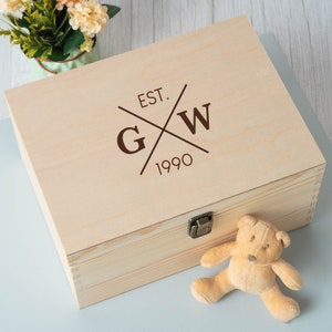 May include: A wooden box with a hinged lid and a brass latch. The lid is engraved with the letters "G" and "W" in a crossed design, along with the text "EST. 1990". A small teddy bear is sitting on a light blue surface next to the box.