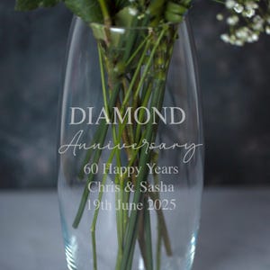 May include: A clear glass vase with the words "DIAMOND Anniversary" and "60 Happy Years" etched on the front. The names "Chris & Sasha" and the date "19th June 2025" are also etched on the vase. The vase contains green stems and white flowers.