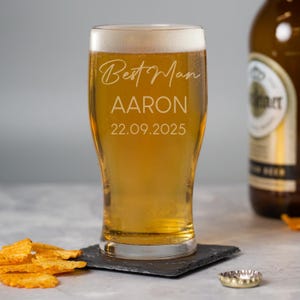 May include: A clear beer glass filled with golden beer and foam. The glass is engraved with "Best Man", the name "Aaron", and the date "22.09.2025". The glass sits on a black coaster, with a bottle of beer in the background.
