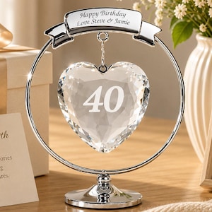 May include: A silver-toned metal stand featuring a circular frame and a base, holding a faceted clear crystal heart ornament. The heart is engraved with the number 40, and the top banner is engraved with the text Happy Birthday Love Steve & Jamie.
