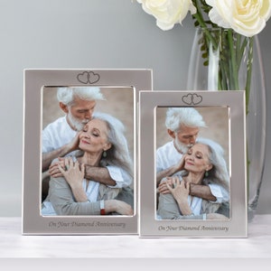 Op de afbeelding: Twee zilveren fotolijsten met een stel in elk. De lijsten hebben een hartvormig ontwerp aan de bovenkant en de tekst "On Your Diamond Anniversary" aan de onderkant.