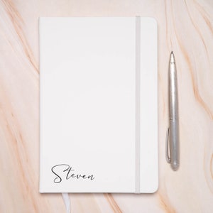 Op de afbeelding: Een wit notitieboek met een zilveren pen en de naam "Steven" op de voorkant.