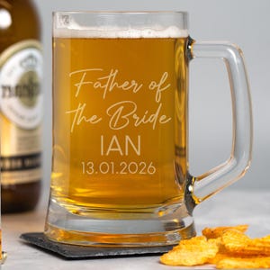 May include: A clear glass beer mug filled with beer, featuring the etched text "Father of the Bride" followed by the name "IAN" and the date "13.01.2026". The mug has a handle and is set on a dark coaster, with potato chips in the foreground.