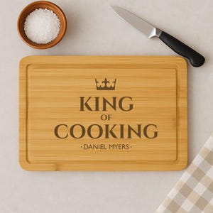 Op de afbeelding: Een lichtbruin bamboe snijplank met de tekst "King of Cooking" en een kroonafbeelding. De naam "Daniel Myers" is onder de tekst gegraveerd.