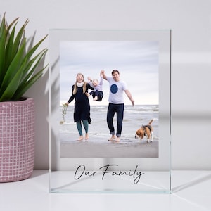 May include: A glass picture frame with a photo of a family of three and their dog on a beach. The frame has the words "Our Family" written on the bottom.