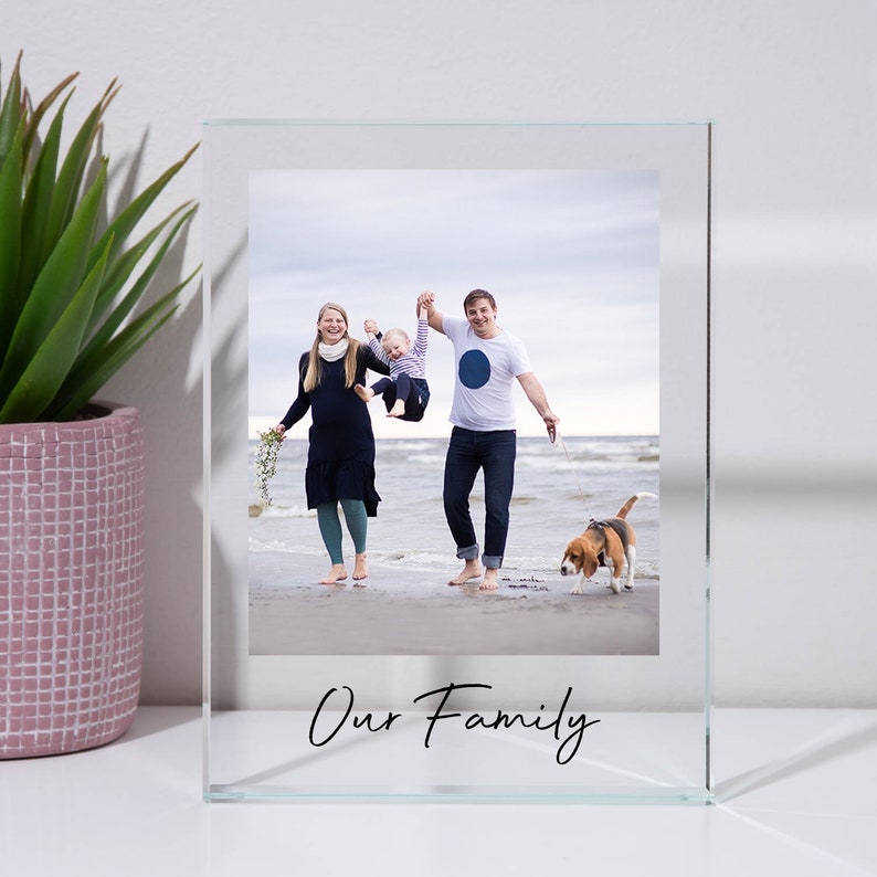 May include: A glass picture frame with a photo of a family of three and their dog on a beach. The frame has the words "Our Family" written on the bottom.