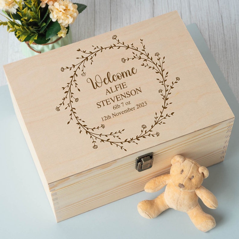 Op de afbeelding: Een houten bewaardoos met een gegraveerd bloemenkransontwerp op de deksel. De krans bevat de tekst "Welcome Alfie Stevenson 6lb 7oz 12th November 2023".