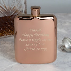 May include: A rose gold hip flask with a matching cap. The flask has rounded corners and engraved text that reads "Daniel Happy Birthday Have a tipple on me Lots of love Charlotte xxx."