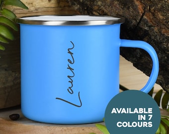 Könnte beinhalten: Eine hellblaue Emaille-Tasse mit weißem Rand und einer schwarzen Aufschrift "Lauren". Die Tasse steht auf einer Holzoberfläche mit grünem Laub im Hintergrund. Der Text "AVAILABLE IN 7 COLOURS" ist in einem blaugrünen Kreis sichtbar.