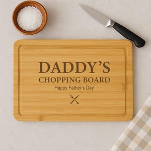 Può includere: Un tagliere rettangolare in bambù marrone con il testo "Daddy's Chopping Board" e "Happy Father's Day" inciso su di esso. Un paio di simboli di coltello e forchetta incrociati sono anche incisi sul tagliere.