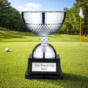 Puede incluir: Un trofeo de golf plateado brillante con una copa texturizada y una base negra. El trofeo está sobre un campo de golf verde, con una pelota y una bandera en el fondo. La base tiene una placa que dice "Any Engraving Here".