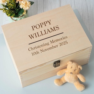 May include: A light-colored wooden memory box with the name "POPPY WILLIAMS" and "Christening Memories 10th November 2025" engraved on the lid. A small, tan teddy bear rests beside the box. A small bouquet of flowers is in the background.