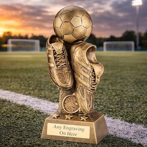 Peut inclure: Trophée de football en bronze avec un ballon et une paire de chaussures. La base rectangulaire porte l'inscription « Any Engraving On Here ». L'arrière-plan montre un terrain de football avec un coucher de soleil.