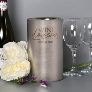 May include: Silver wine cooler with the text "Wine Lovers Emma & Ross" engraved on the side. The cooler is next to two wine glasses and white flowers.