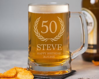 May include: A clear glass beer mug filled with beer, featuring the number 50 inside a laurel wreath, the name "STEVE", and "HAPPY BIRTHDAY 26.07.2025" etched on the front. The mug has a handle and sits on a black coaster. Potato chips and a bottle cap are in the foreground.