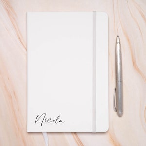 Op de afbeelding: Een wit notitieboek met een zilveren pen en de naam "Nicola" op de voorkant.