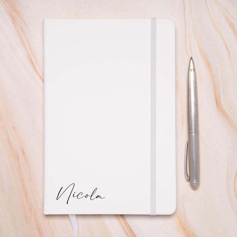 Op de afbeelding: Een wit notitieboek met een zilveren pen en de naam "Nicola" op de voorkant.